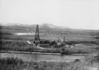 The Dingman No. 1 well on the bank of the Sheep River with the Calgary Petroleum Products office in the distance. This is probably before the well hit wet gas on May 14, 1914, as there are no signs of pipelines running from the well and no storage tanks at the site. In later years, the plant would develop on the flat land to the west of the derrick, in the middle distance in this photo. <br />Source: Glenbow Archives, NB-16-604