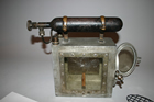 This canary cage has an oxygen tank attached to the top of it, to revive the canary should it passed out from inhaling poisonous carbon monoxide gas. The wooden stick inside is a perch on which the canary rested, or from which it fell if it was poisoned.